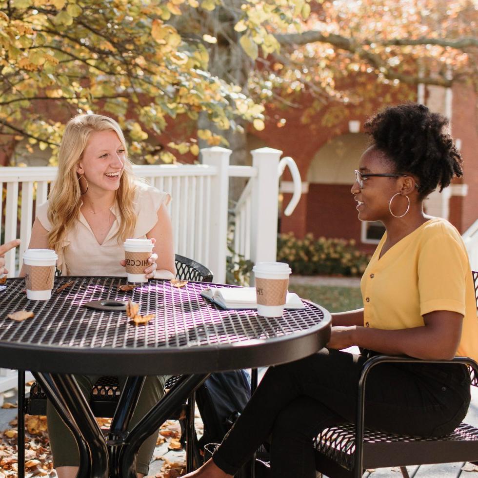 博彩app推荐(艾斯拜瑞大学市)校园里的学生们坐在桌子旁愉快地与朋友聊天，桌子上放着咖啡杯.